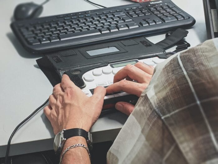 Hands reading an electronic braille display that's in front of a computer keyboard