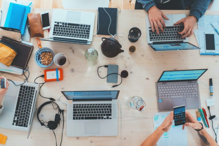 People sitting at a table with laptops, phones, headphones, chargers, and snacks.